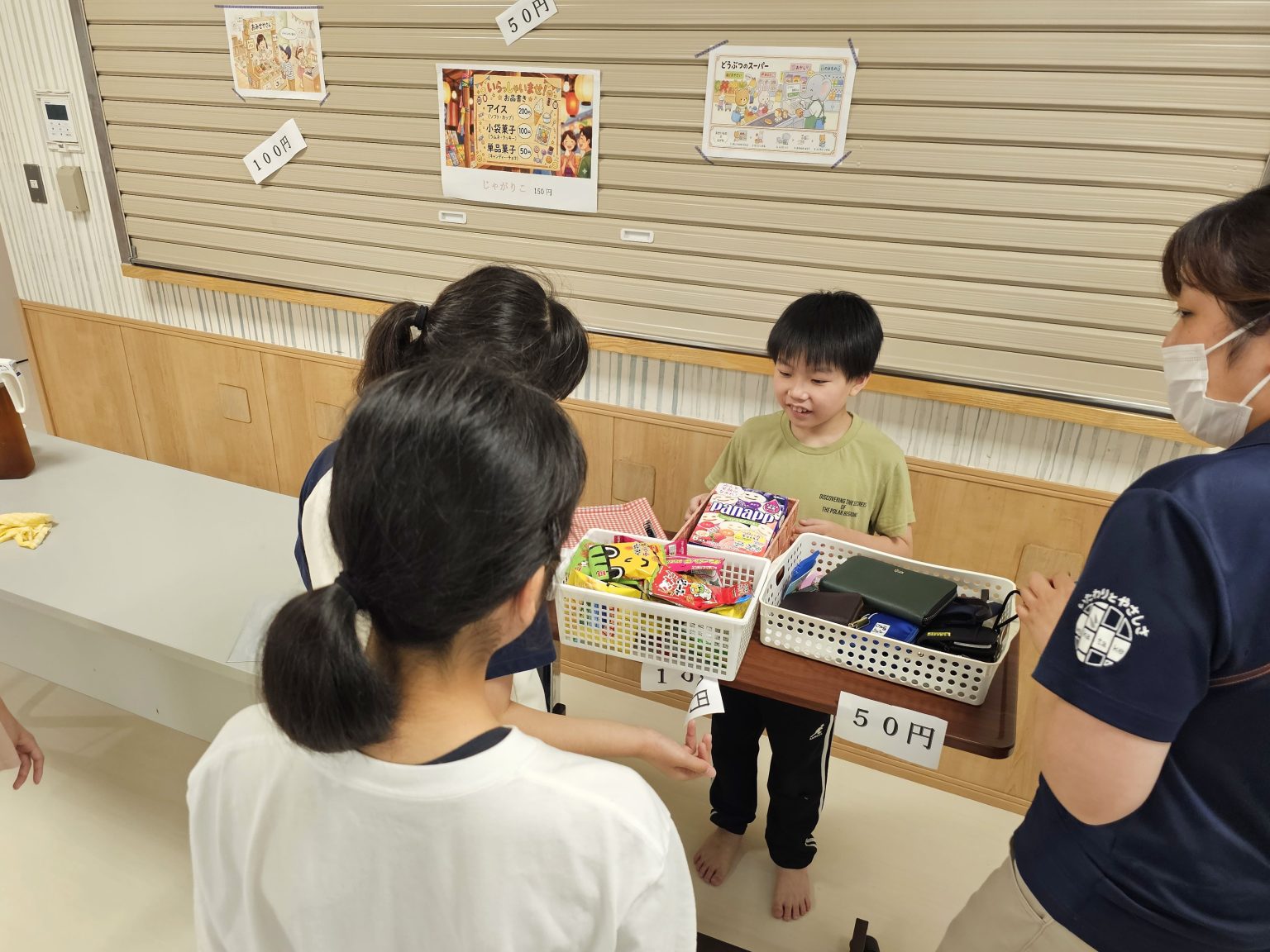 児童が職員と一緒にお店屋さんごっこの買い物体験をしている様子。教育的な活動風景。